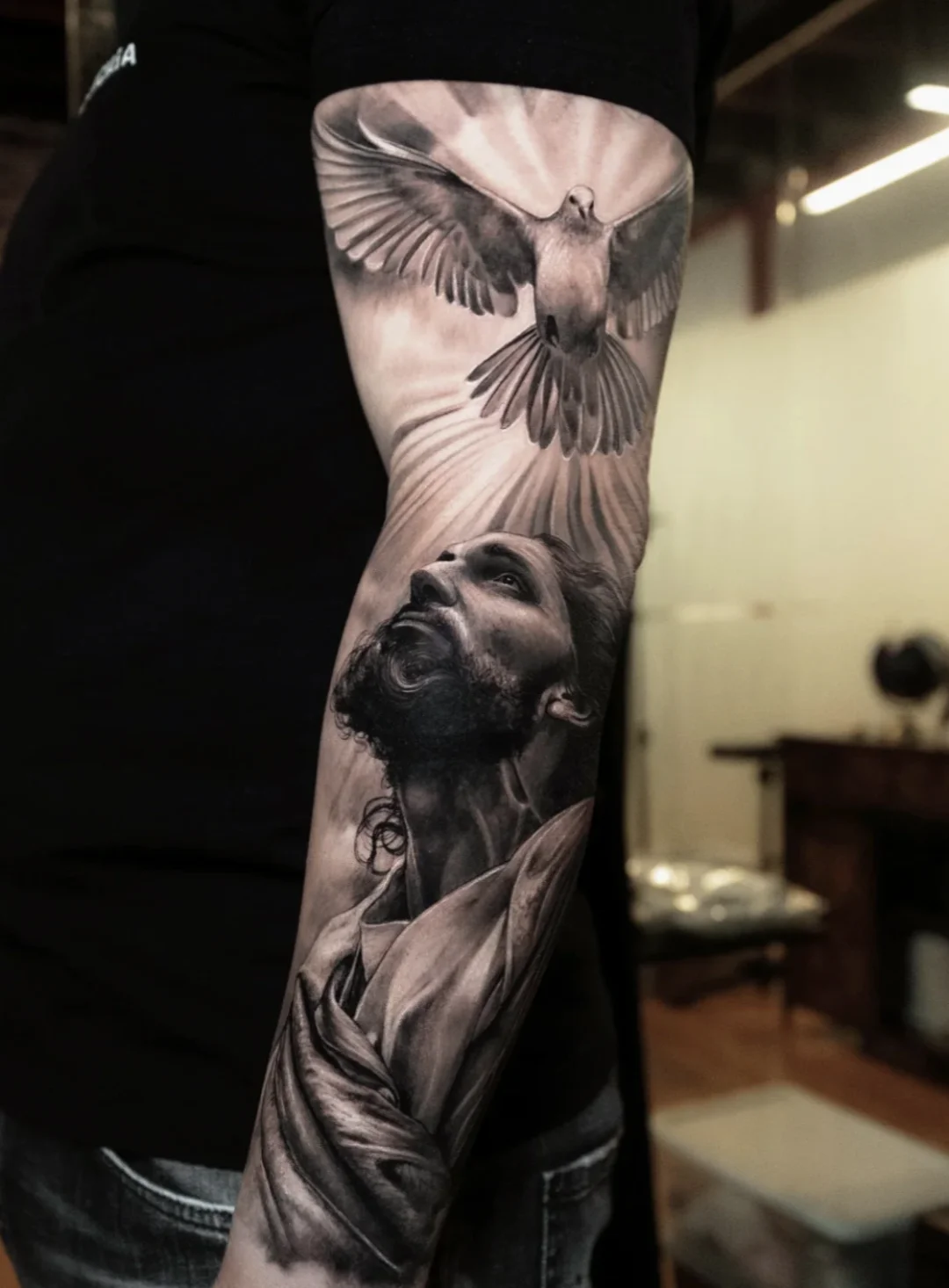 This highly detailed arm piece depicts Jesus looking upwards toward a radiant dove, using smooth black and gray shading for a powerful, life
